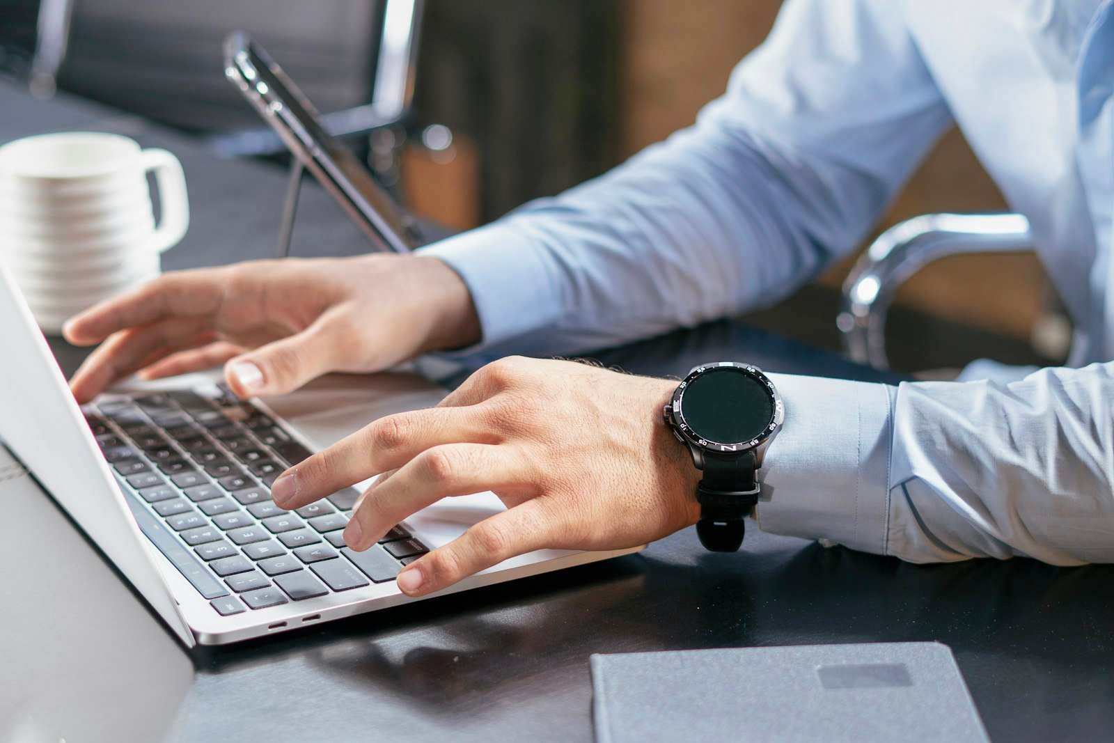Close-up of a professional in a light blue shirt typing on a laptop at a desk, with a phone on a stand and a white mug beside it