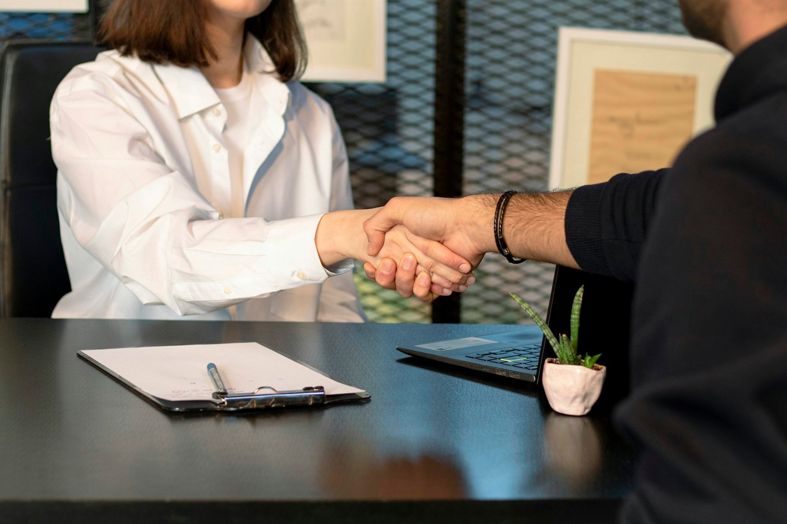 A woman in a white shirt and a man in a dark sweater shaking hands across a desk with a clipboard and a laptop